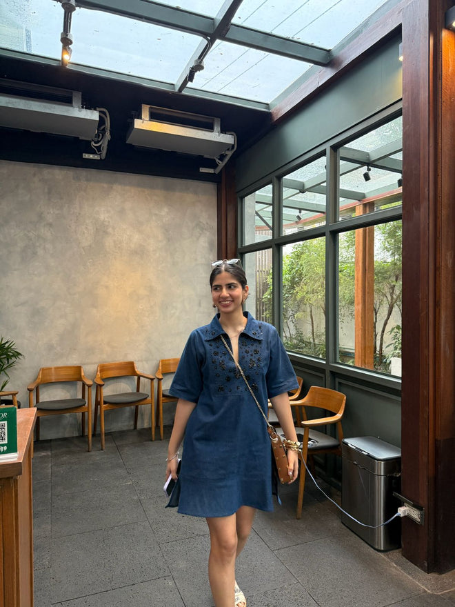 Woman in a blue dress standing in a modern indoor setting with large windows.
