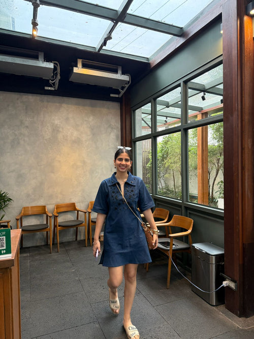 Woman in a blue denim hand-embroidered 
dress standing in a modern indoor setting with large windows.